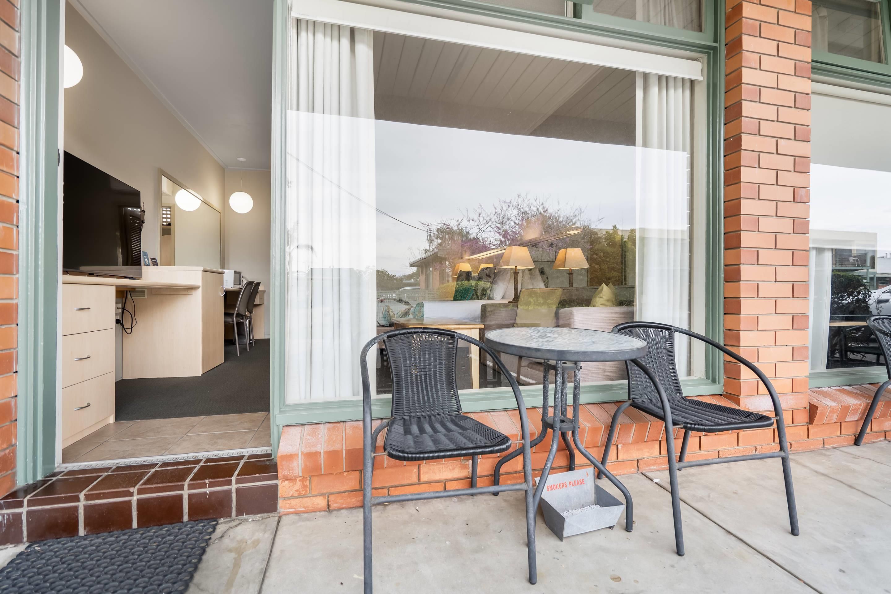 An outdoor seating area with two chairs and a table, a brick wall, and a window with blinds.