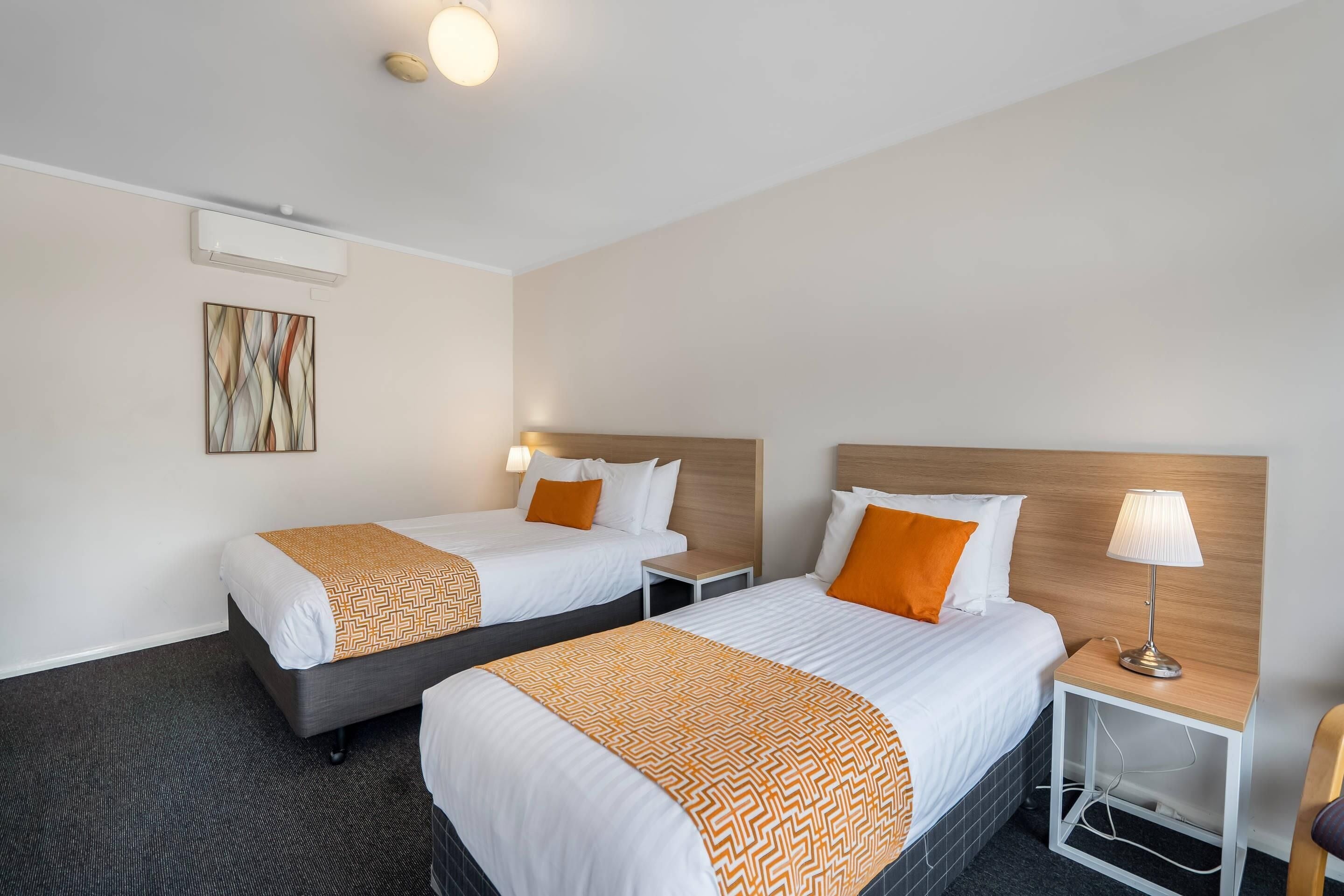 A hotel room with two beds, a bedside table with a lamp, a wall-mounted air conditioner, and a painting.