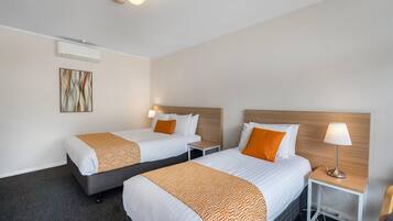 A hotel room with two beds, a bedside table with a lamp, a wall-mounted air conditioner, and a painting.