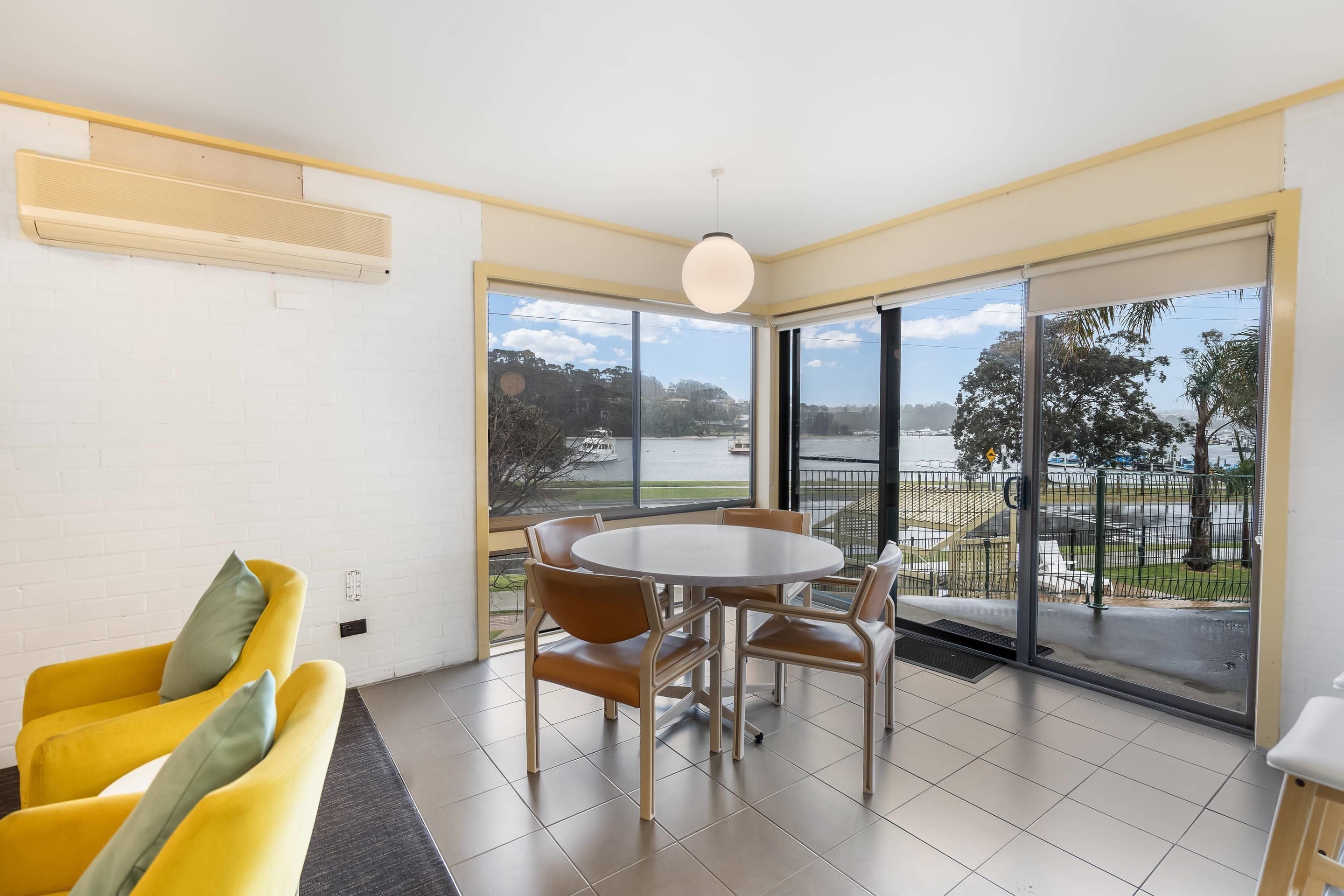 A modern room with a dining table and chairs, a yellow sofa, and large windows offering a view of a body of water and greenery.
