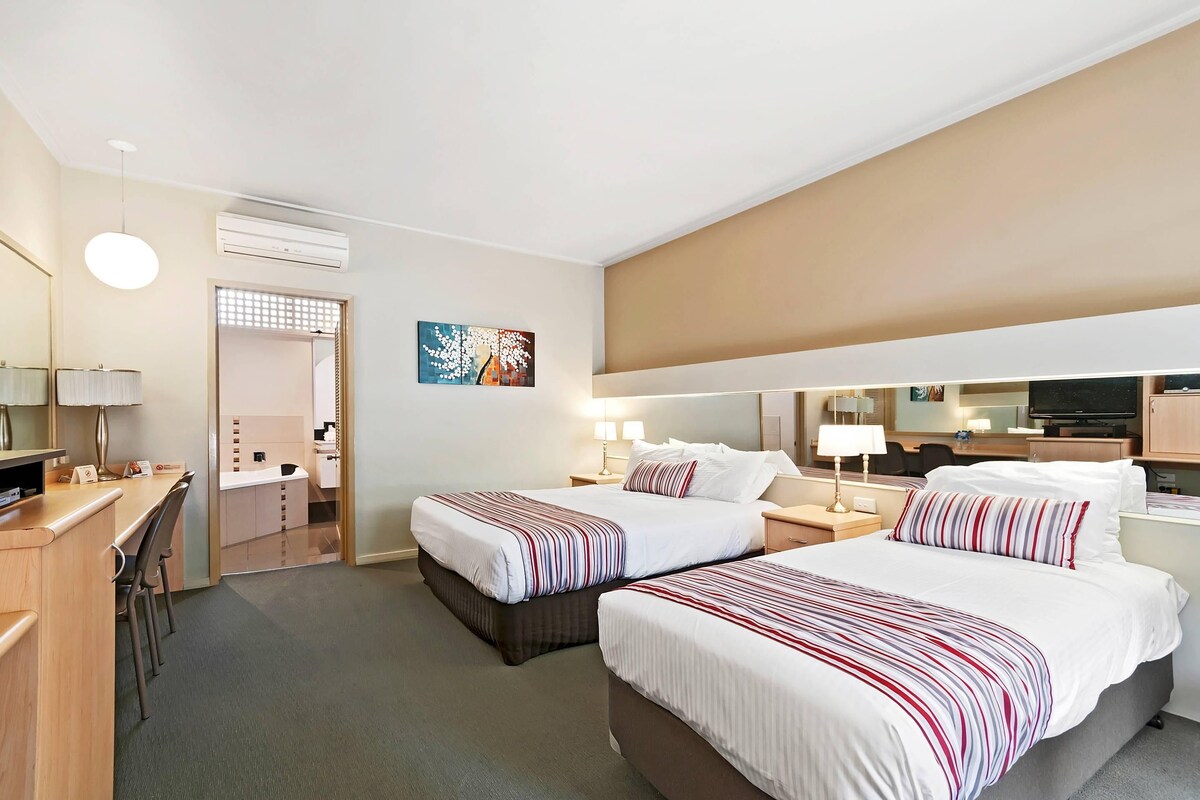 A hotel room with two beds, a desk, a chair, a TV, and a bathroom visible through an open door.