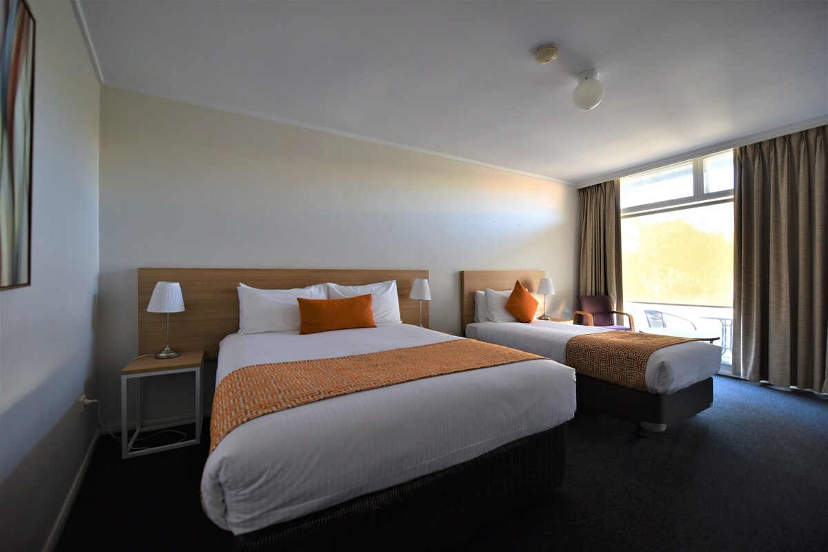 A hotel room with two beds, a nightstand with a lamp, a window with curtains, and a view of the outdoors.