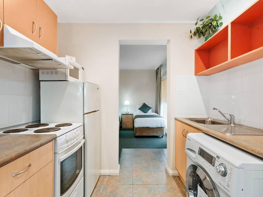 A compact kitchen with a stove, oven, and washing machine, leading to a bedroom with a bed and nightstand.