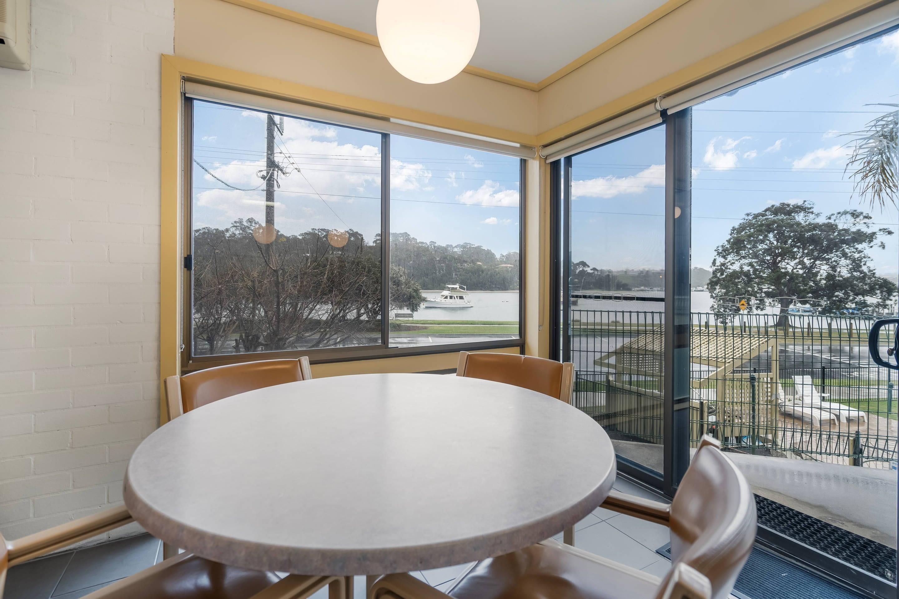 A room with a round table and four chairs, large windows with a view of a body of water and trees, and a balcony with a railing.