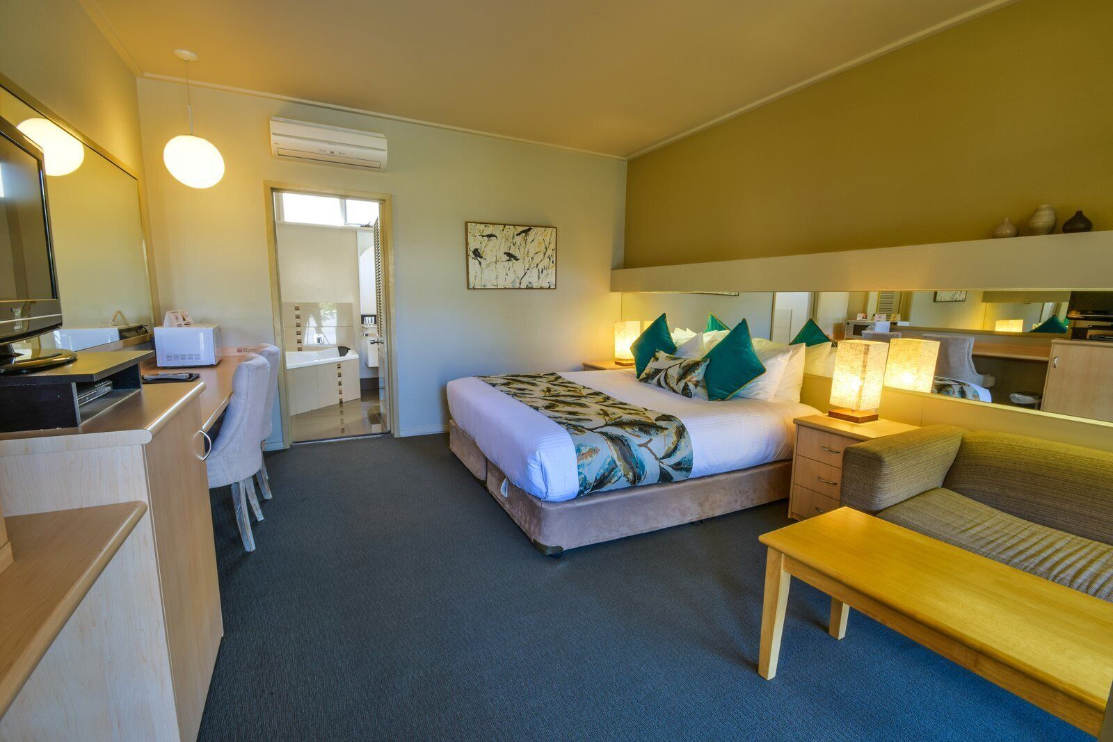 A hotel room with a bed, bedside table, lamp, desk, chair, and a bathroom visible through an open door.