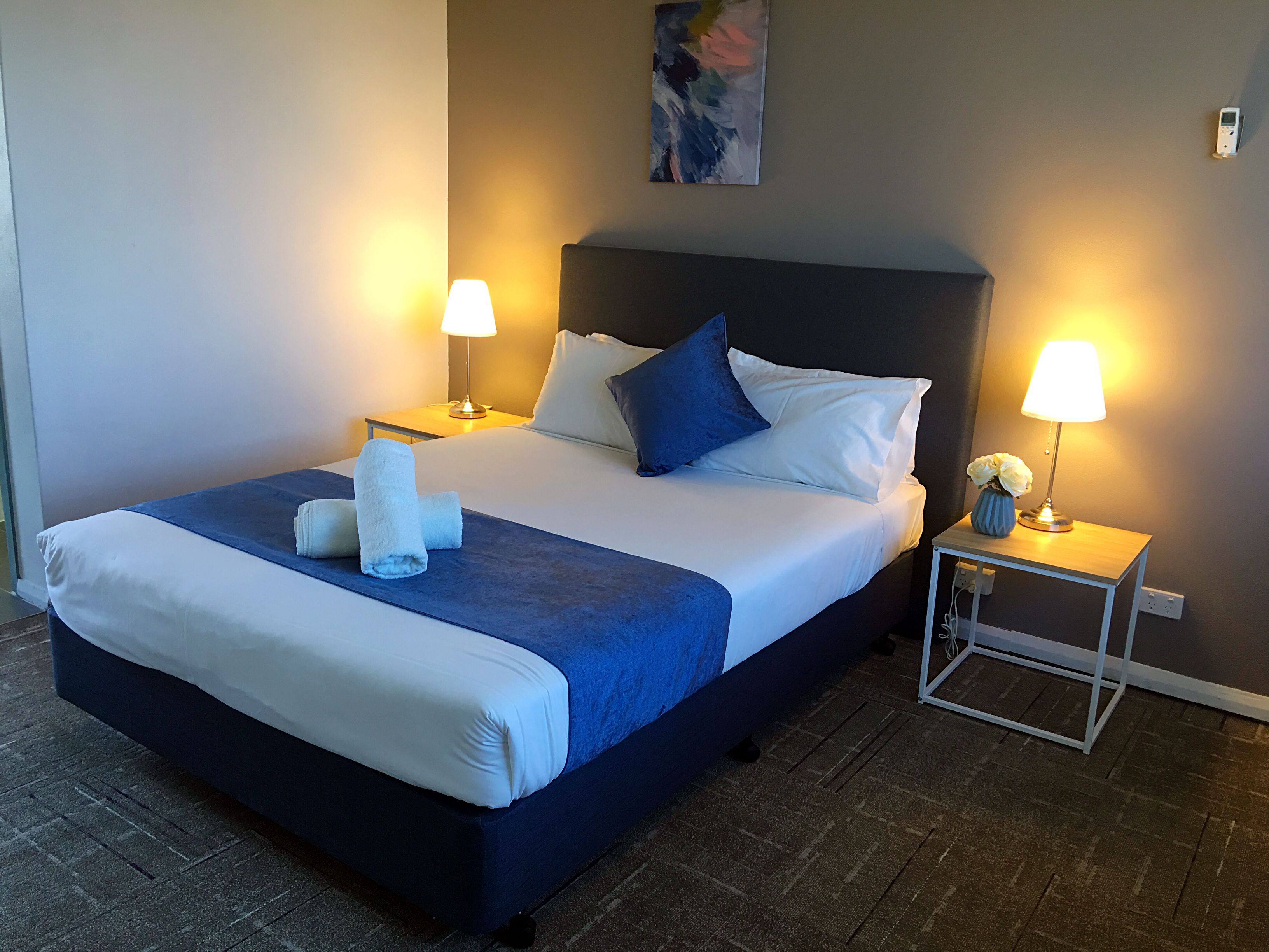 A neatly made bed with white and blue bedding, two bedside lamps, and a small vase with flowers on a nightstand.
