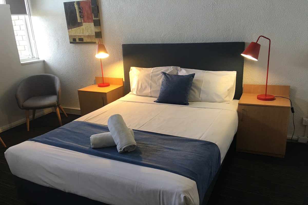 A neatly made bed with a blue and white duvet, two pillows, and a rolled towel. There are two bedside tables with lamps, a chair, and a wall-mounted artwork.