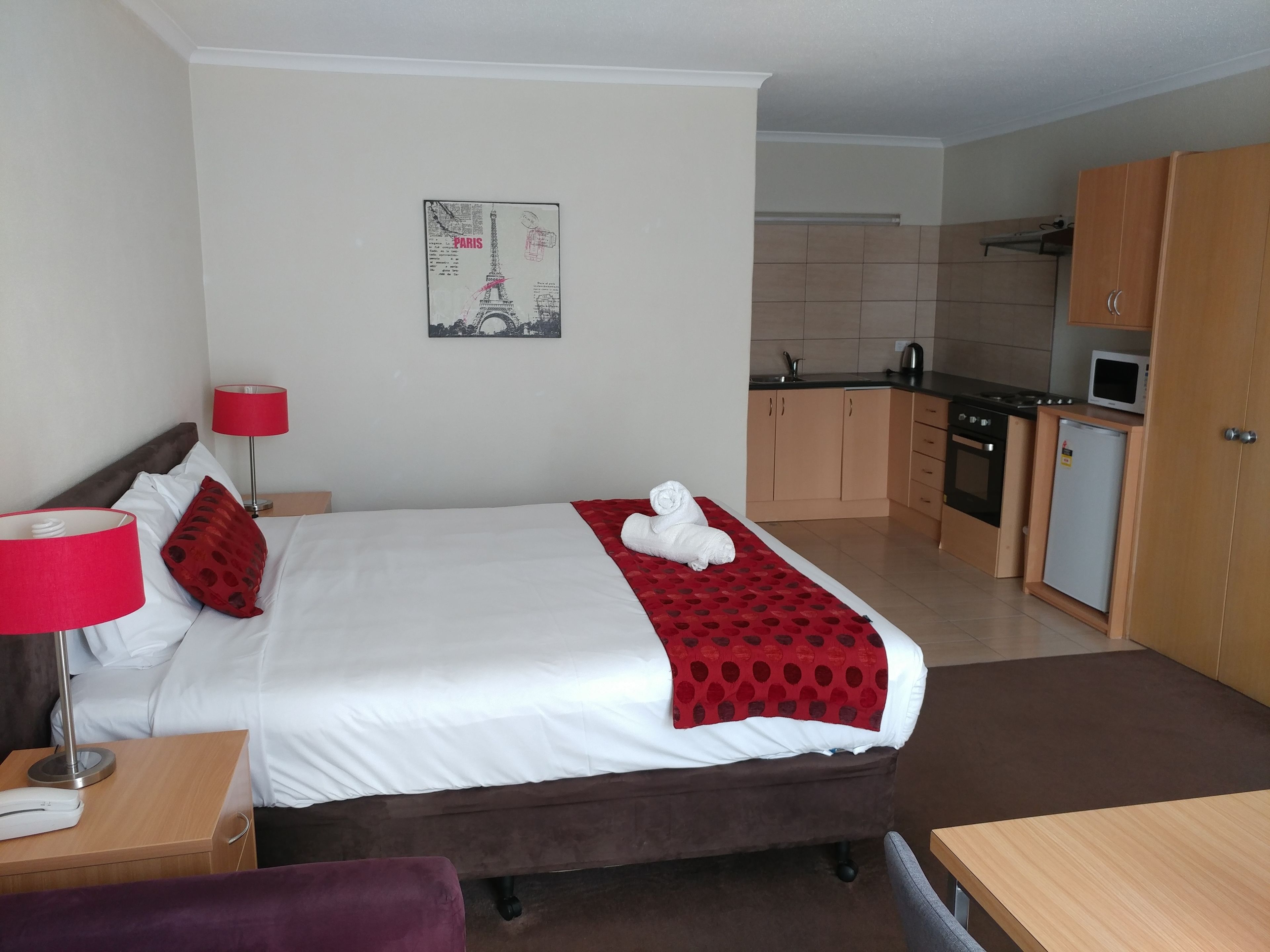 A hotel room with a bed, a nightstand, a desk, a chair, a red lamp, a red and white patterned blanket, a towel, a kitchen area with cabinets, a stove, a microwave, and a refrigerator.