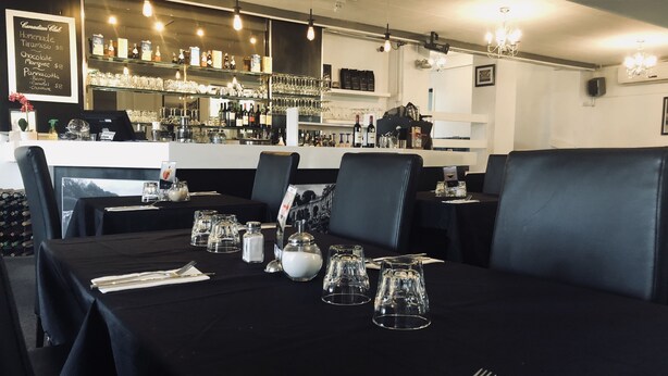 Un restaurante con manteles negros, mesas dispuestas con esmero y una zona de bar al fondo.