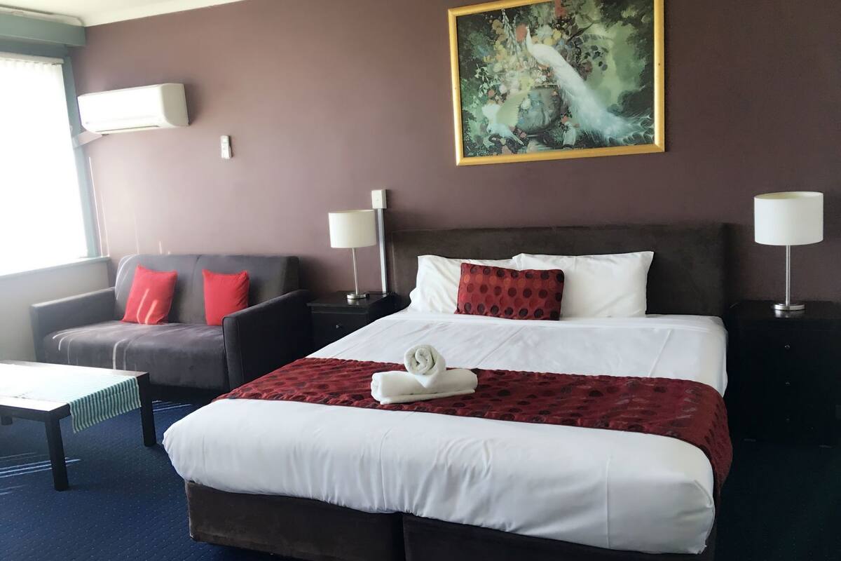 A hotel room with a bed, a sofa, two nightstands, a painting, and a window.