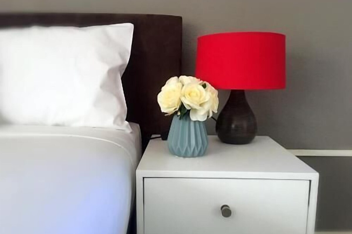 A bed with a white pillow and a dark headboard, a small white nightstand with a red lamp and a vase of flowers, and a white drawer.
