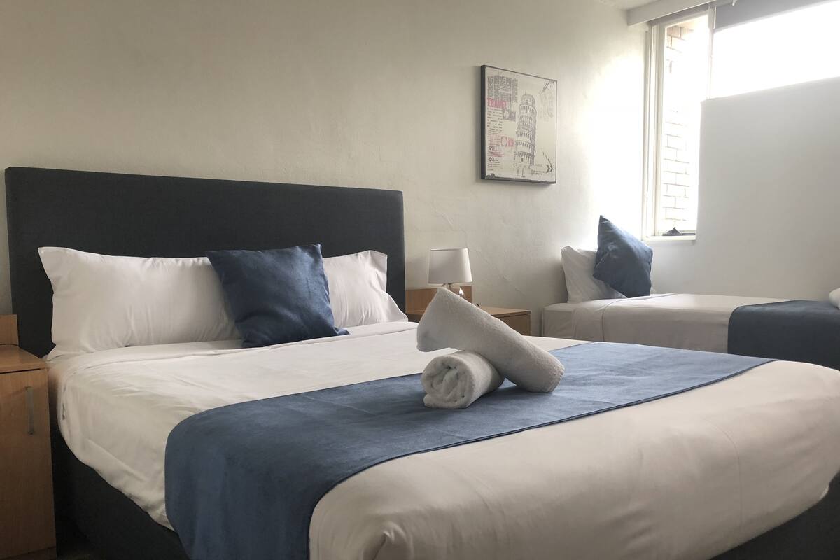 A hotel room with a large bed, two bedside tables, a nightstand, a lamp, and a window with blinds.