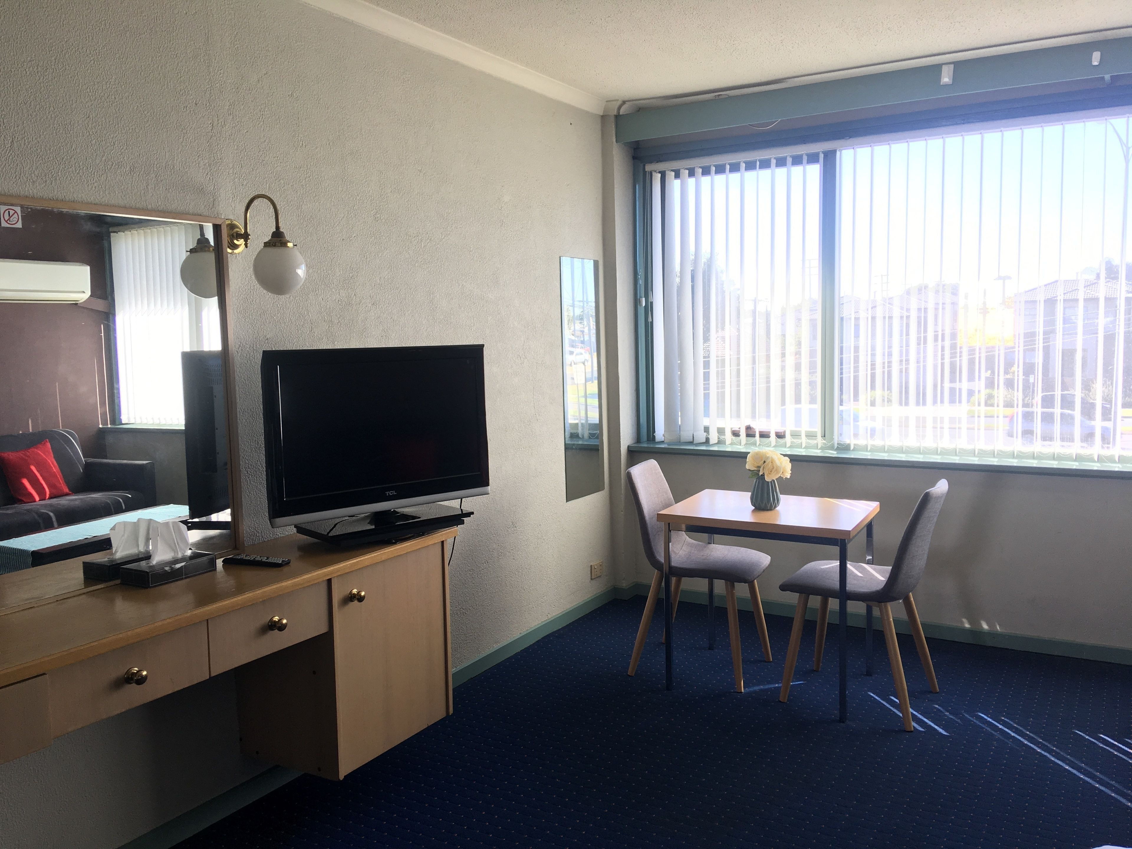 A hotel room with a desk, a flat-screen TV, a dining area with a table and chairs, a window with blinds, and a mirror.