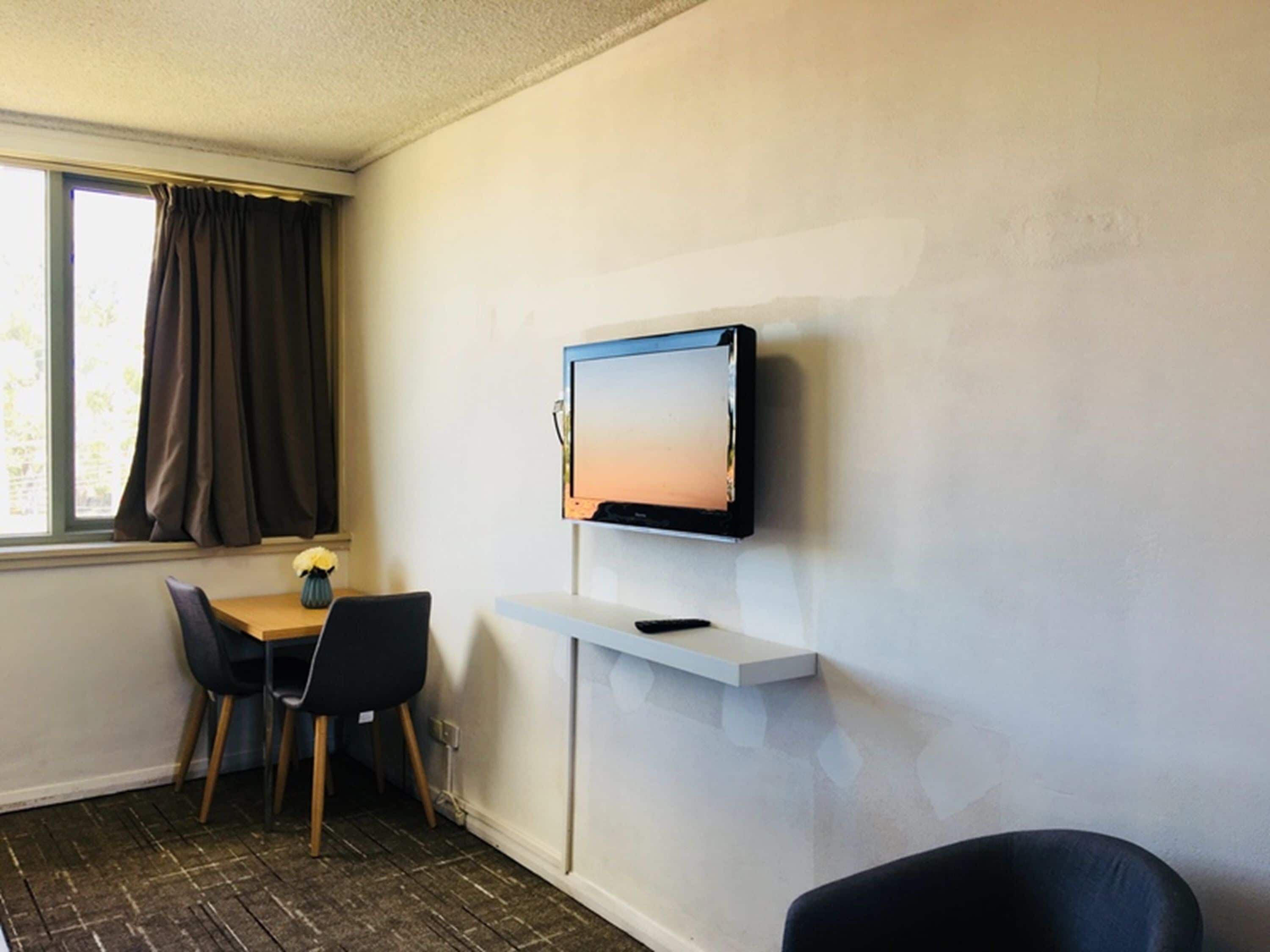 A room with a TV mounted on the wall, a small table with a vase of flowers, two chairs, and a window with curtains.
