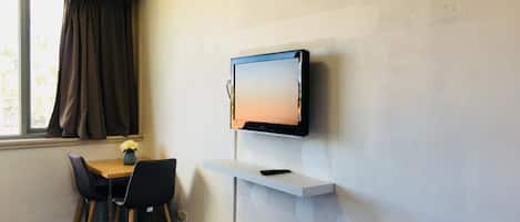 A room with a TV mounted on the wall, a small table with a vase of flowers, two chairs, and a window with curtains.