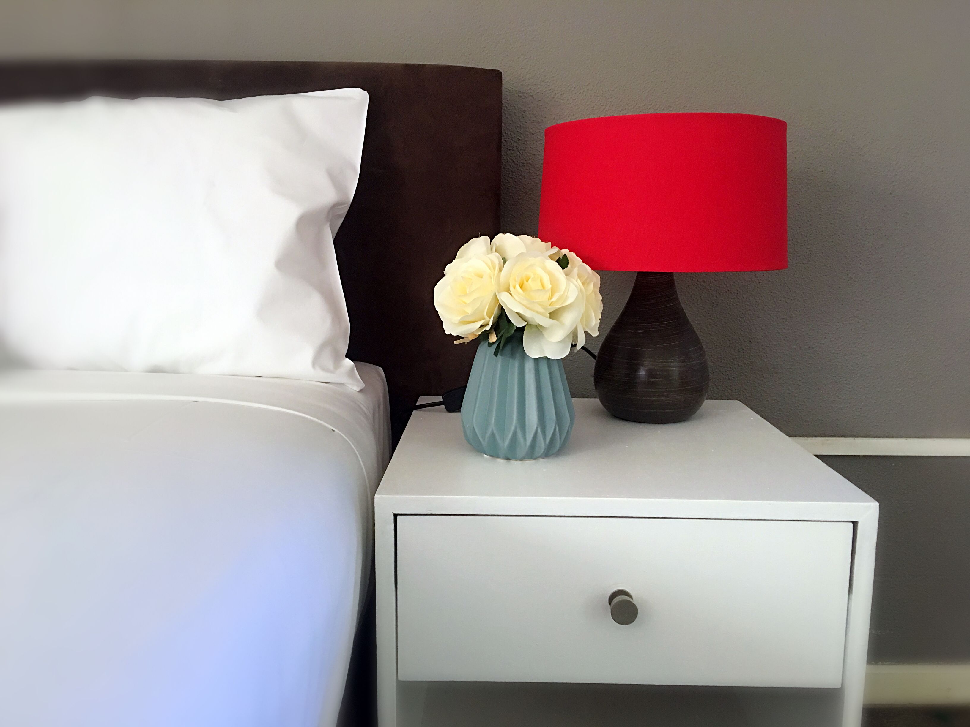 Una cama bien hecha con ropa de cama blanca, un cabecero marrón oscuro, una almohada blanca, una lámpara roja y un florero con flores blancas sobre una mesita de noche blanca.