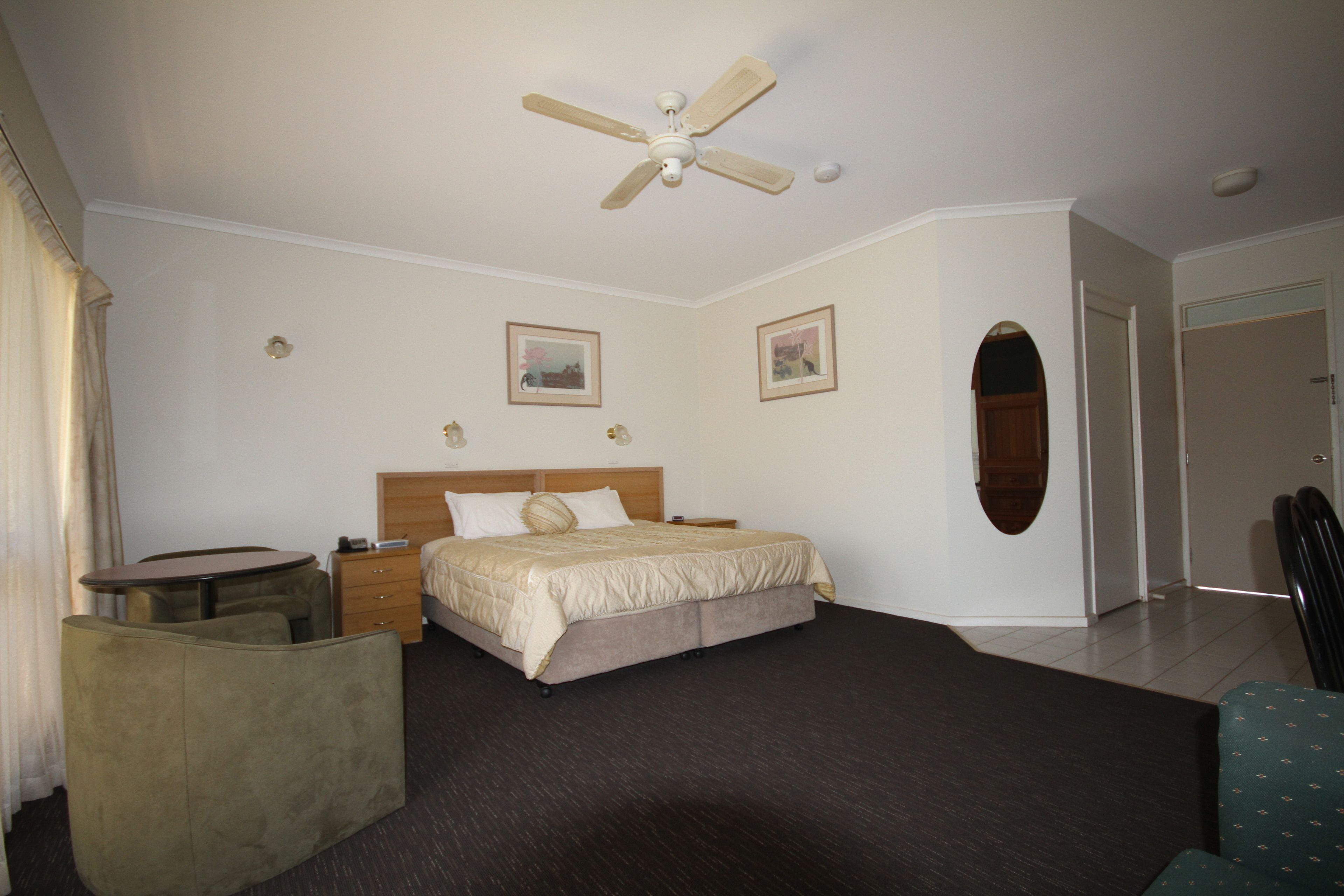Quarto de hotel com cama, escrivaninha, cadeira, ventoinha no teto e duas imagens emolduradas na parede.