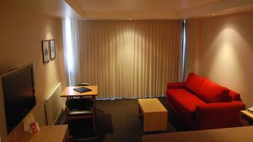 A hotel room with a red sofa, a wooden table, a TV, and a window with curtains.