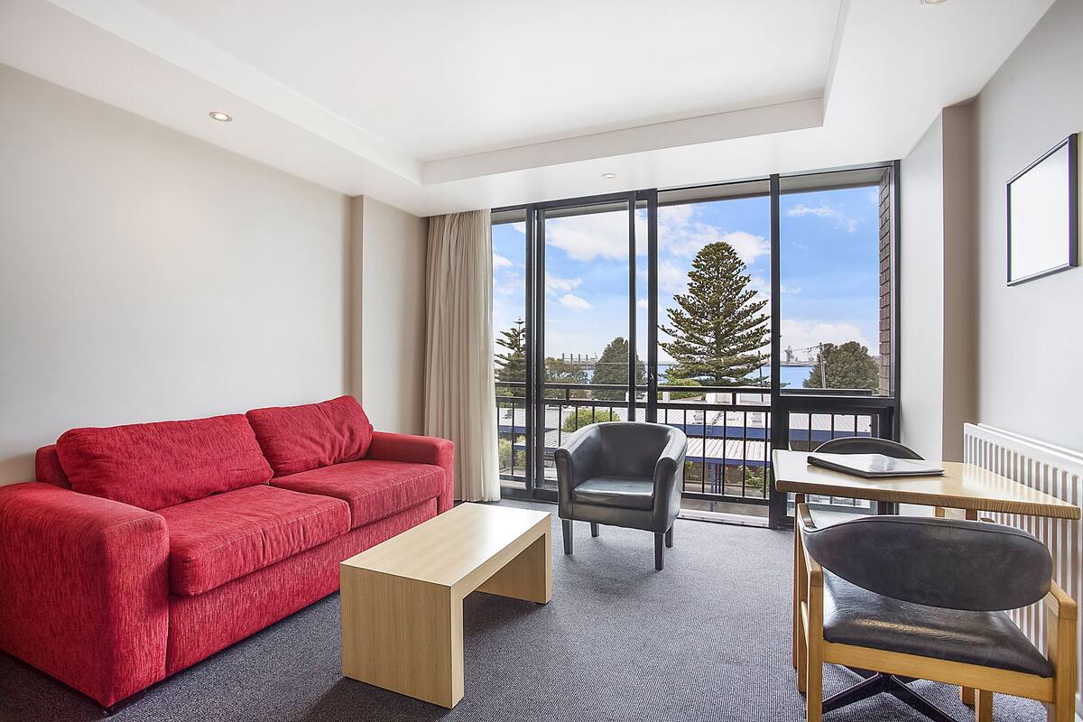 A modern hotel room with a red sofa, a wooden coffee table, a black leather chair, and a view of the outdoors through large windows.