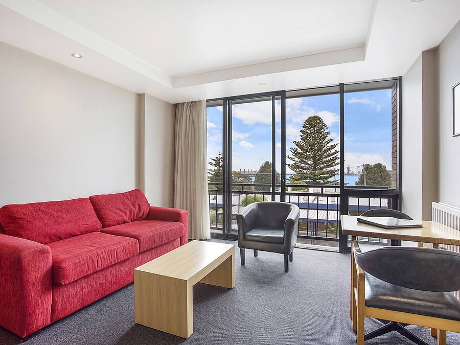 A modern hotel room with a red sofa, a wooden coffee table, a black leather chair, and a view of the outdoors through large windows.