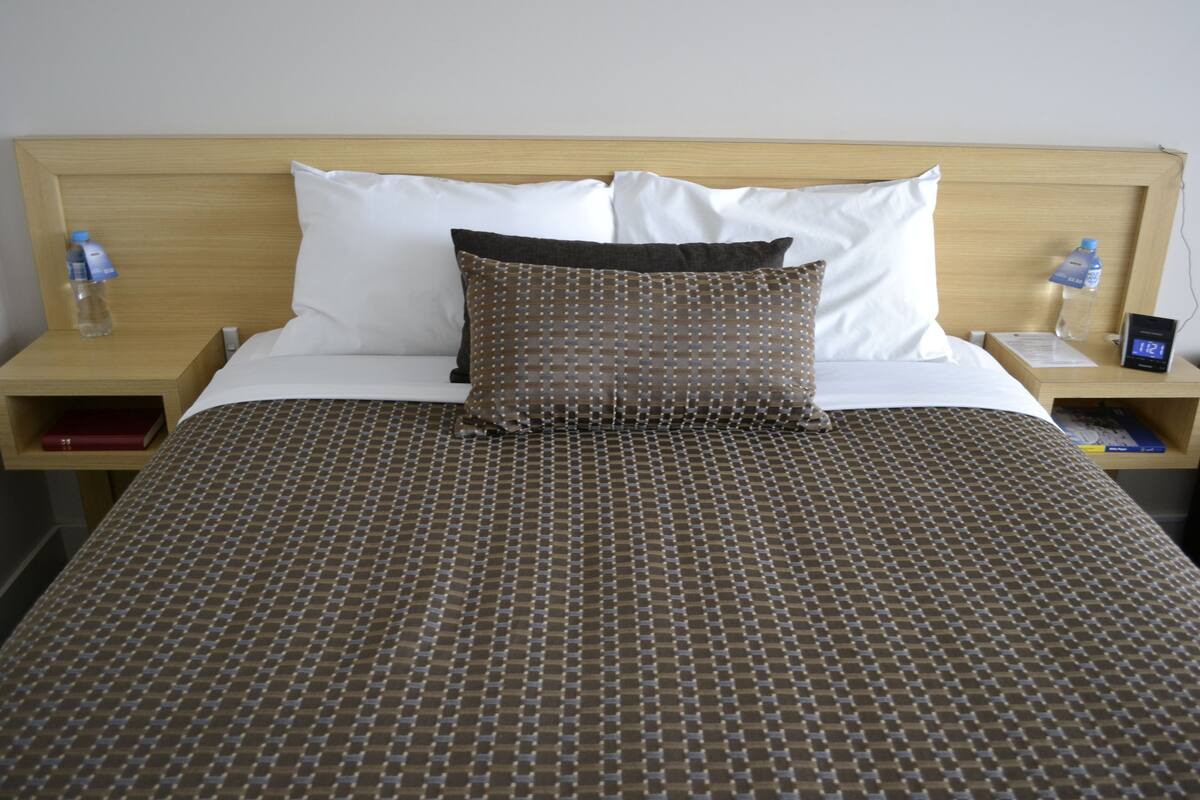 A neatly made bed with a patterned bedspread, two pillows, and a wooden headboard. There are two bedside tables with water bottles and a clock.
