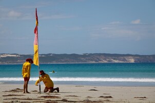 A beach scene with two people in yellow jackets and red hats, one standing and one kneeling, near a flagpole.