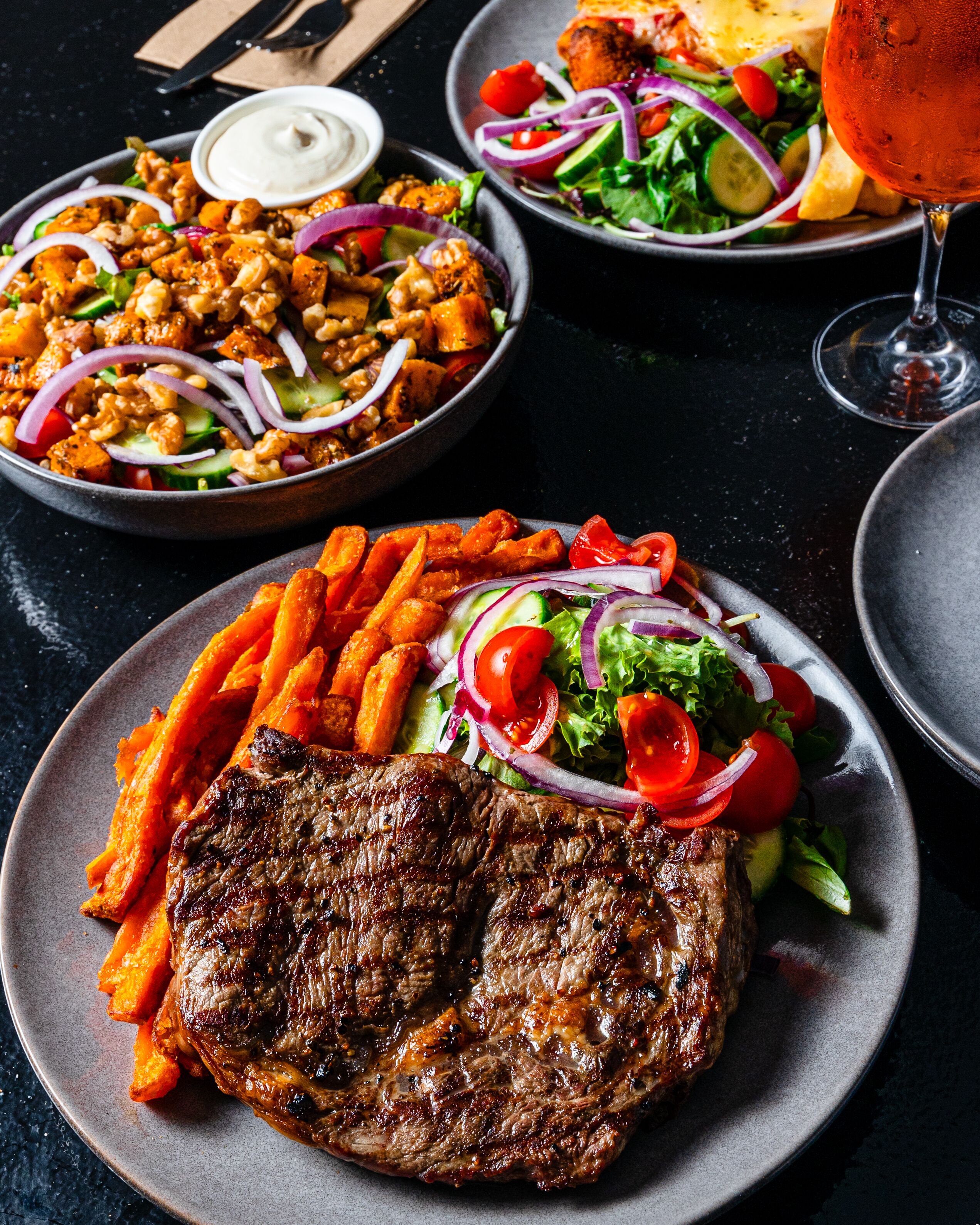 Un plato con un filete a la parrilla, papas dulces fritas y una ensalada de tomates y cebollas.