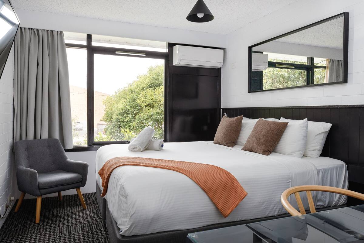 Habitación de hotel con cama, silla, mesa, ventana con cortinas, aire acondicionado y espejo.
