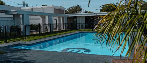 A swimming pool with a tiled deck, surrounded by a fence and a building with a flat roof.
