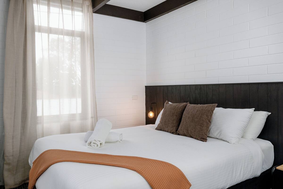Una cama bien hecha con sábanas blancas, un plaid marrón y dos almohadas.
