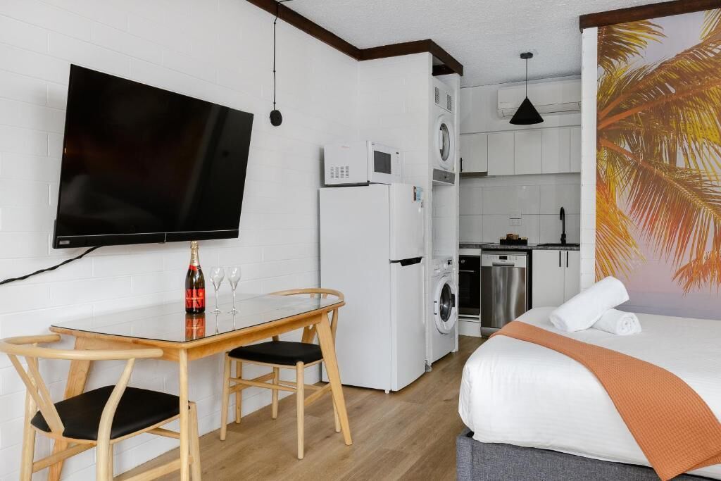 A compact hotel room with a bed, a TV, a dining table with chairs, a washing machine, and a kitchen area.