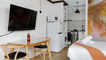 A compact hotel room with a bed, a TV, a dining table with chairs, a washing machine, and a kitchen area.