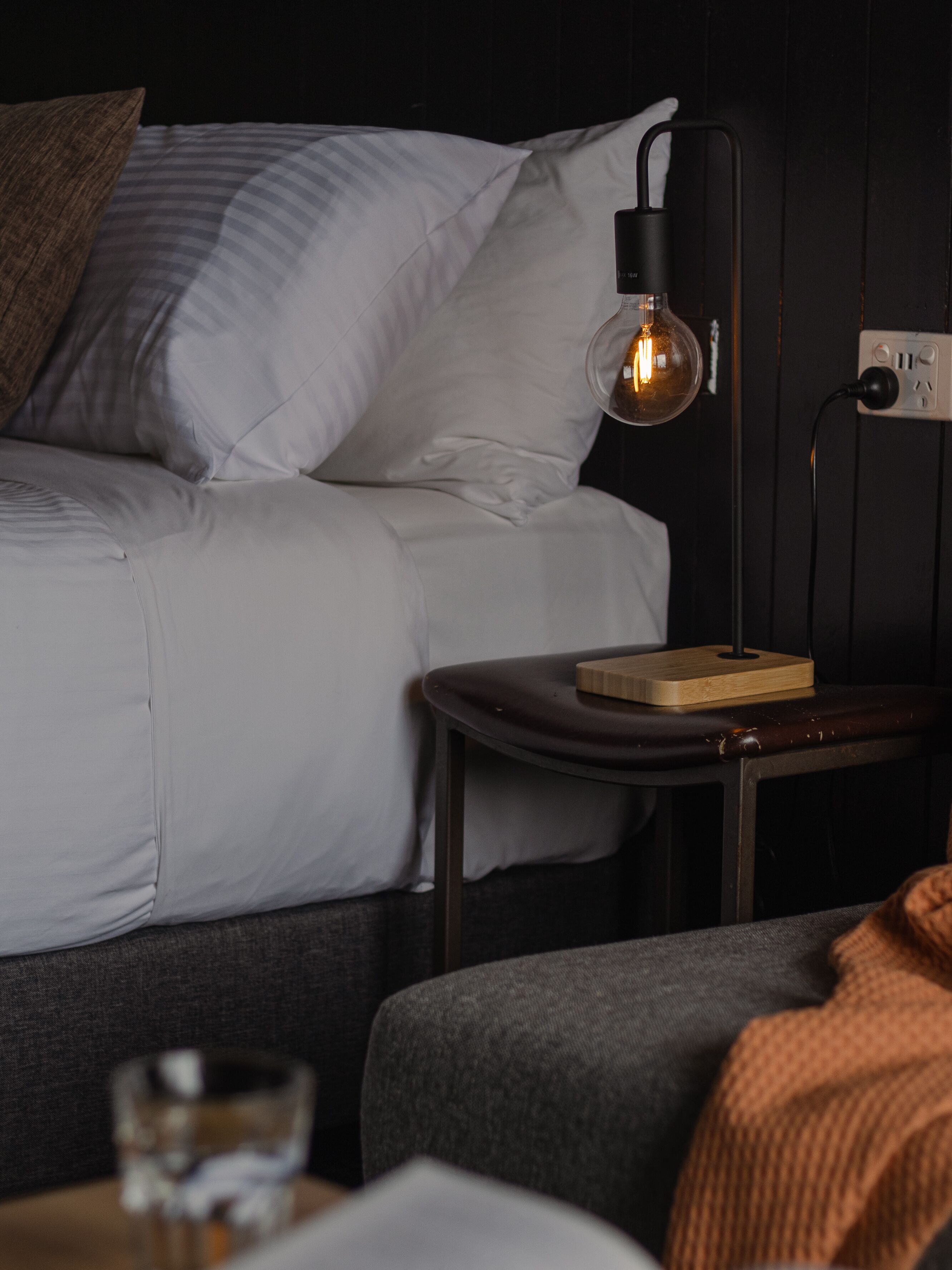 Una cama bien tendida con ropa blanca y una sola almohada marrón. Una mesa moderna con una lámpara y un libro está colocada junto a la cama.
