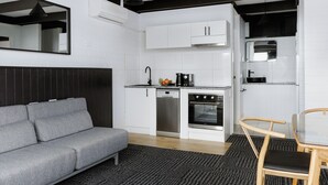 A compact kitchen with white cabinets, a dishwasher, an oven, and a microwave. There's a gray sofa and a small table with two chairs. - m. hotel (Sale)