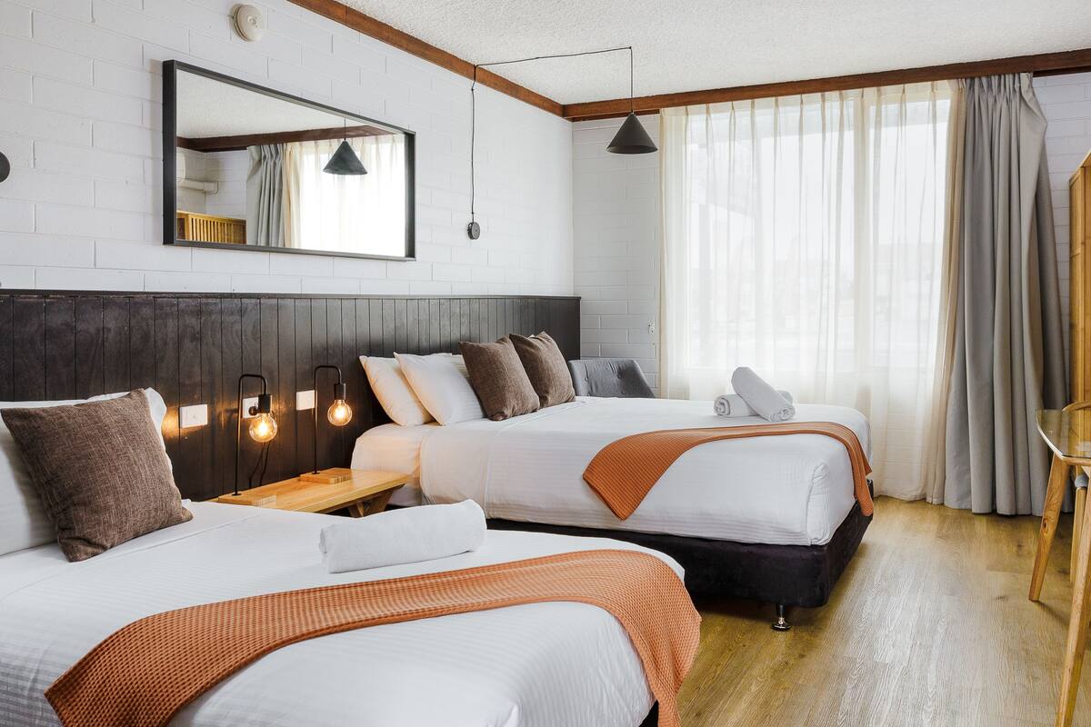 Habitación de hotel con dos camas, cabecero de madera, un espejo grande y una ventana con cortinas.