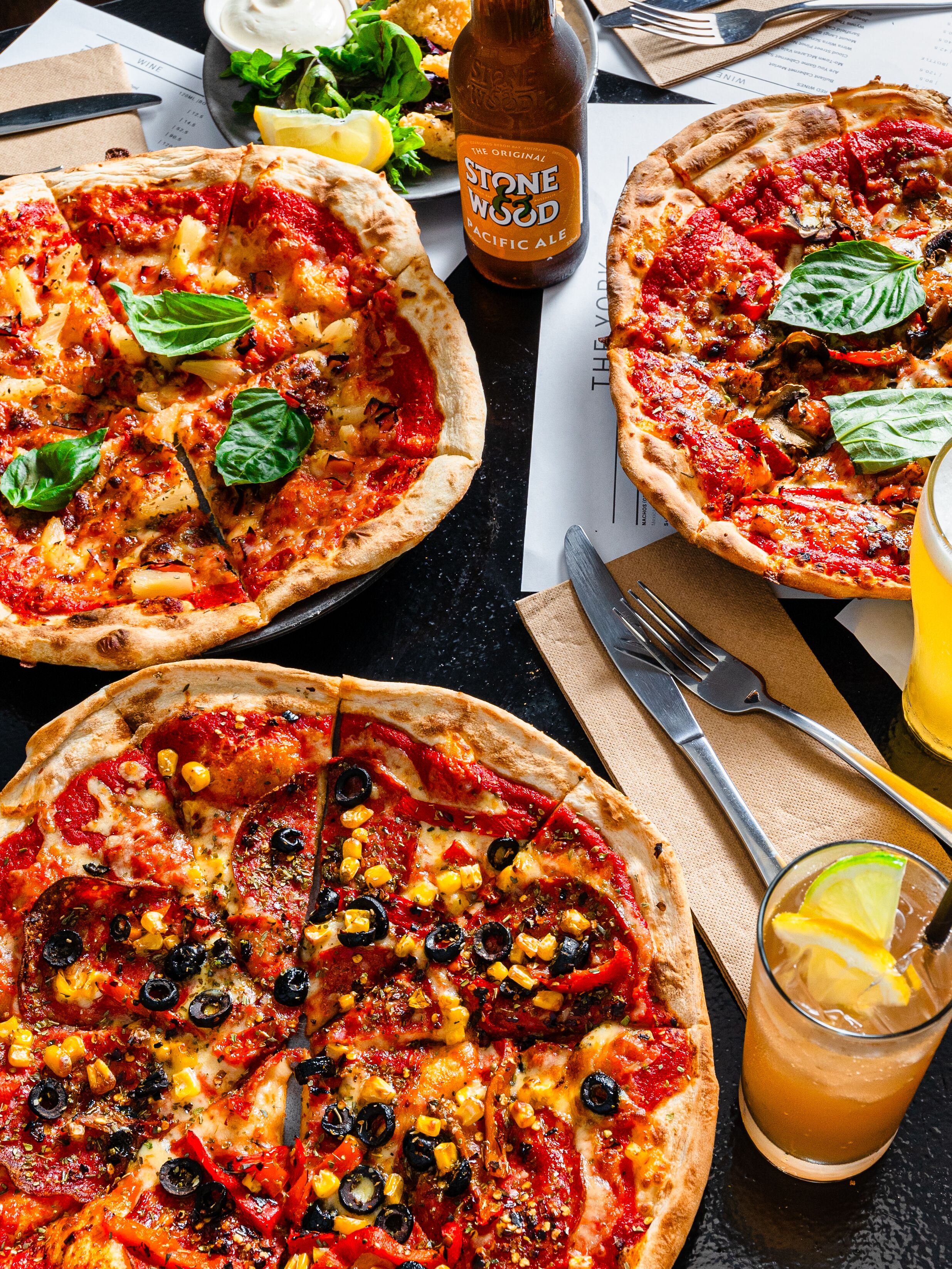 Three pizzas with various toppings, a bottle of Stone Wood Pacific Ale, and a glass of iced tea with lemon.