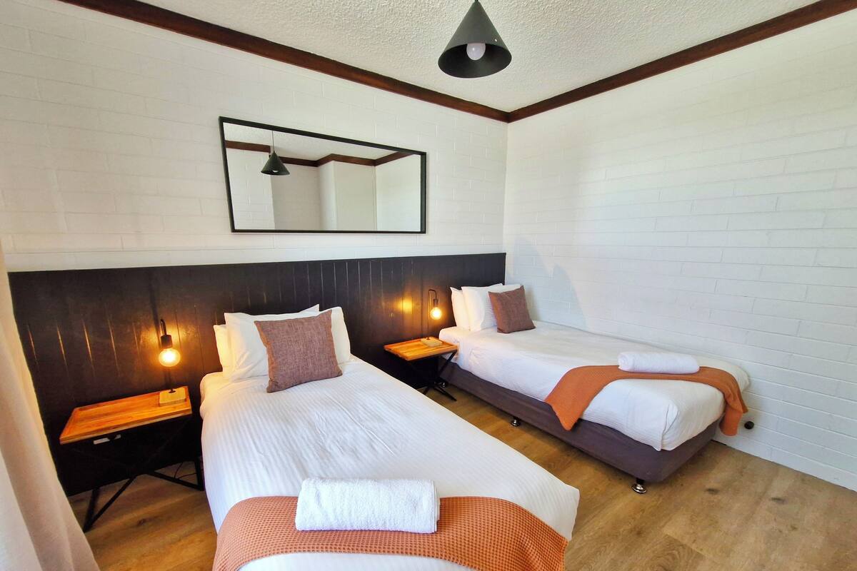 Habitación de hotel con dos camas, cabeceros de madera, un espejo, lámparas de noche y piso de madera.