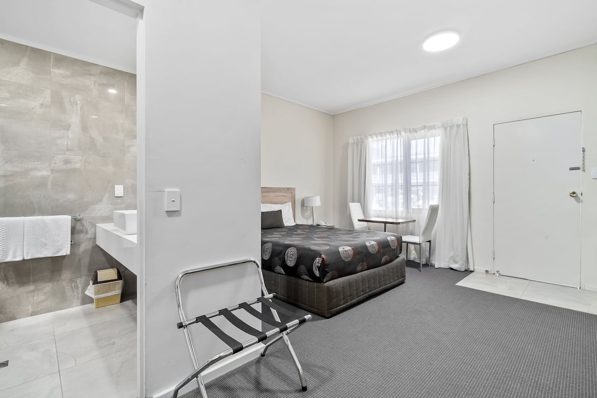 A hotel room with a bed, a chair, a desk, a window with curtains, and a door.
