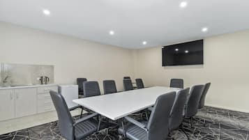 Uma sala de conferências com mesa branca e cadeiras pretas, uma pequena copa e uma televisão grande na parede.