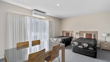 Habitación de hotel con una mesa de vidrio, dos sillas de madera, una cama con ropa de cama estampada, una mesita de noche con una lámpara y un equipo de aire acondicionado.