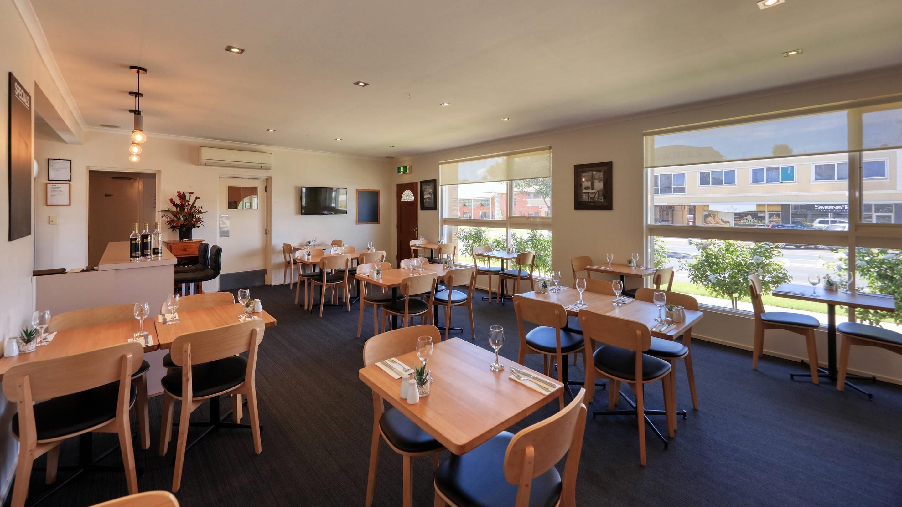 A restaurant with multiple tables and chairs, a flat-screen TV mounted on the wall, and large windows offering a view of the street.