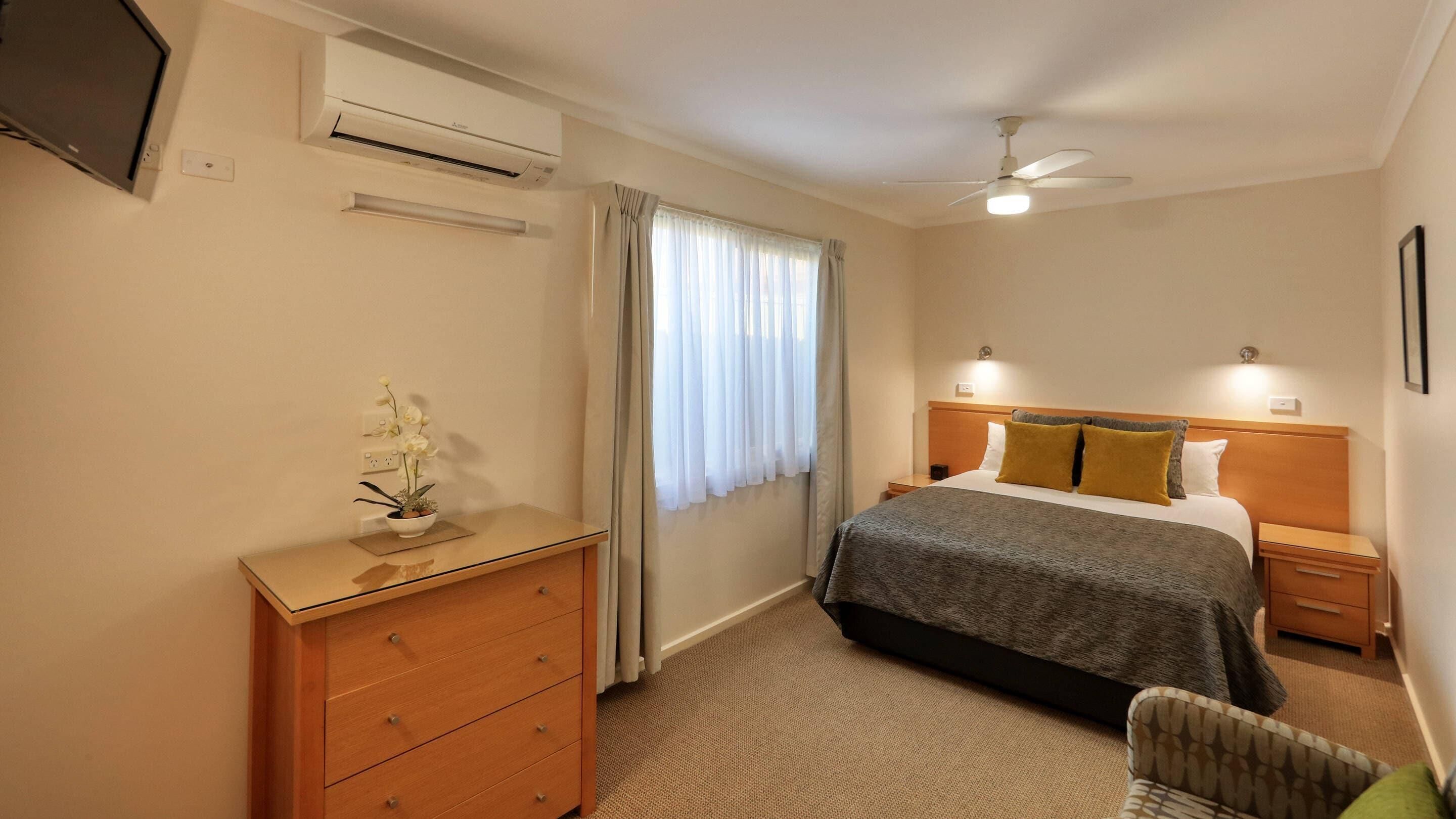 A hotel room with a bed, a nightstand, a dresser, an air conditioning unit, a ceiling fan, and a window with curtains.