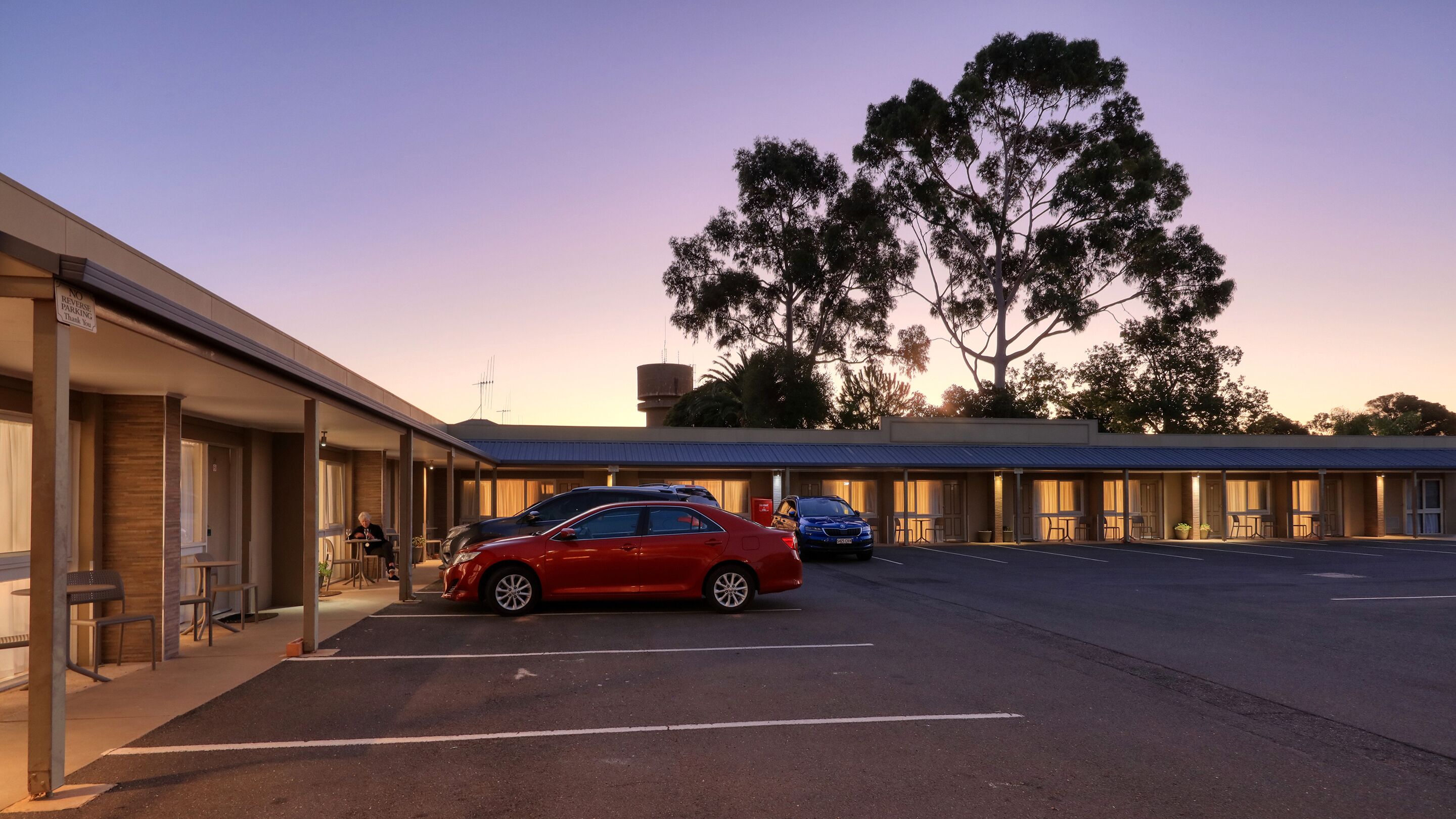 Um motel com um carro vermelho estacionado no estacionamento, uma árvore grande ao fundo e uma placa indicando proibido estacionar em marcha à ré.