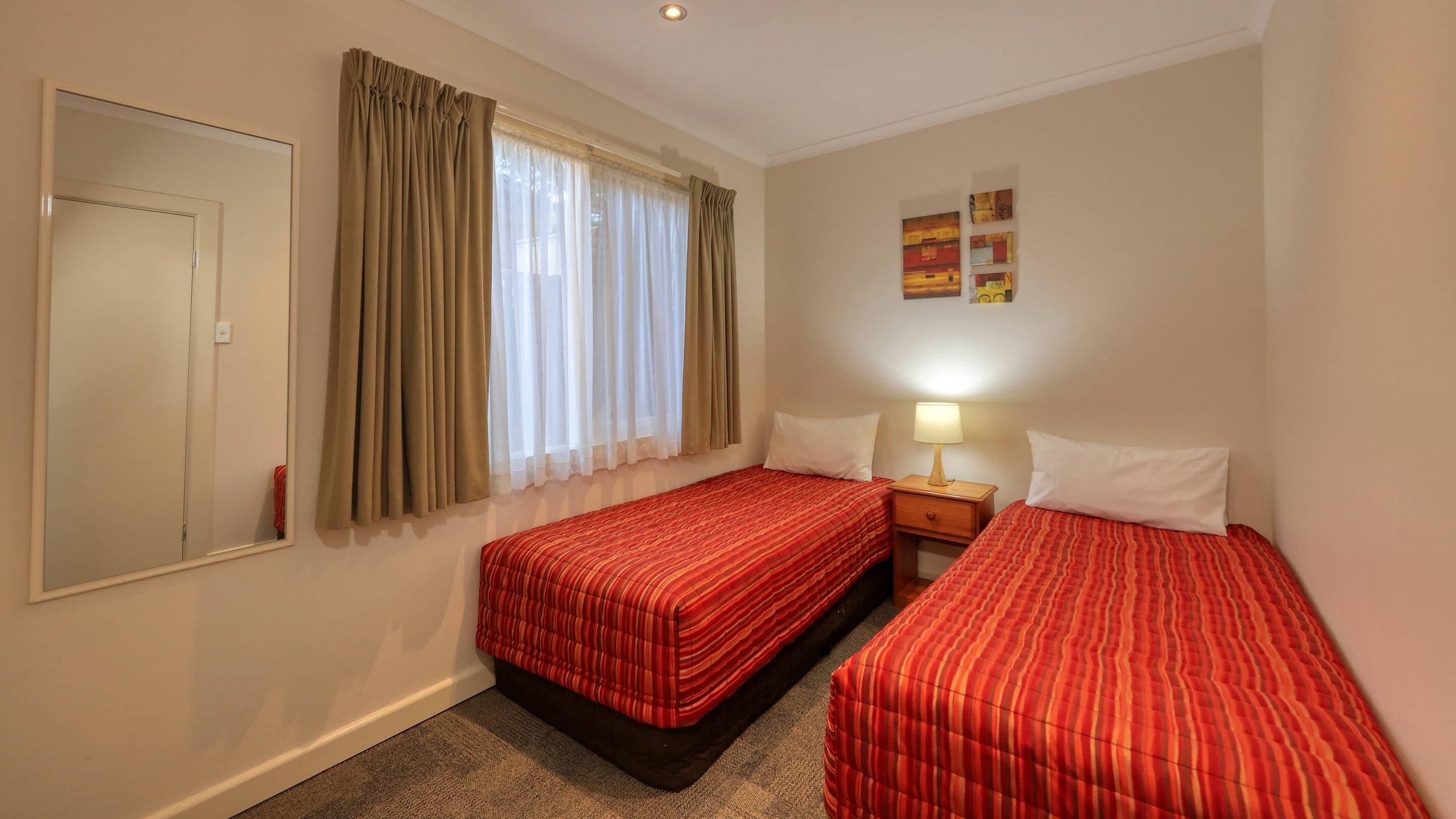 Habitación de hotel con dos camas individuales, una mesita de noche con lámpara, una ventana con cortinas y un espejo en la pared.