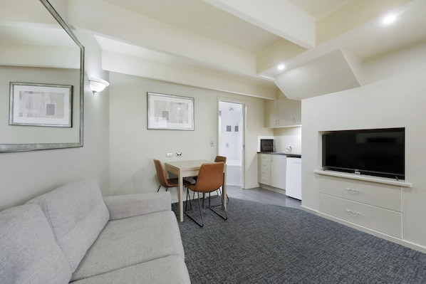 A modern living room with a grey sofa, a dining table with chairs, a flat-screen TV, and a kitchen area with cabinets and a microwave.