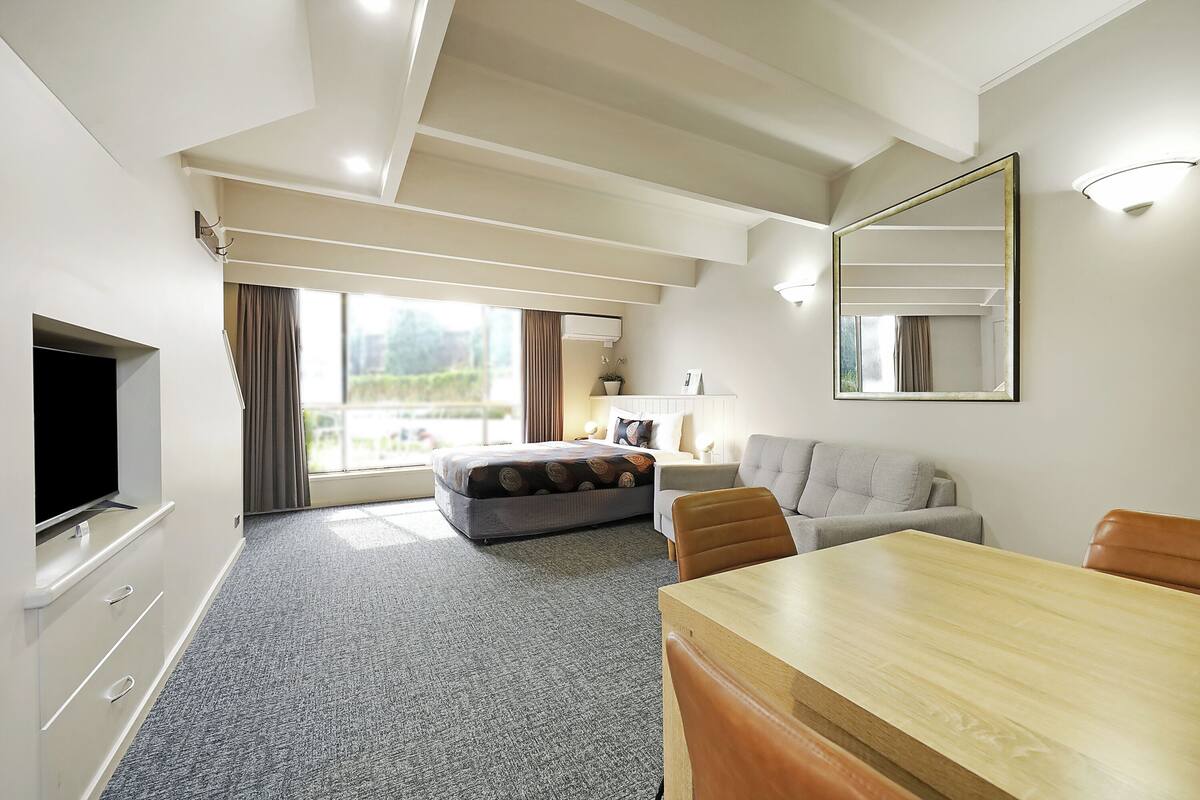 A modern hotel room with a large window, a flat-screen TV, a sofa, a dining table, and a mirror.
