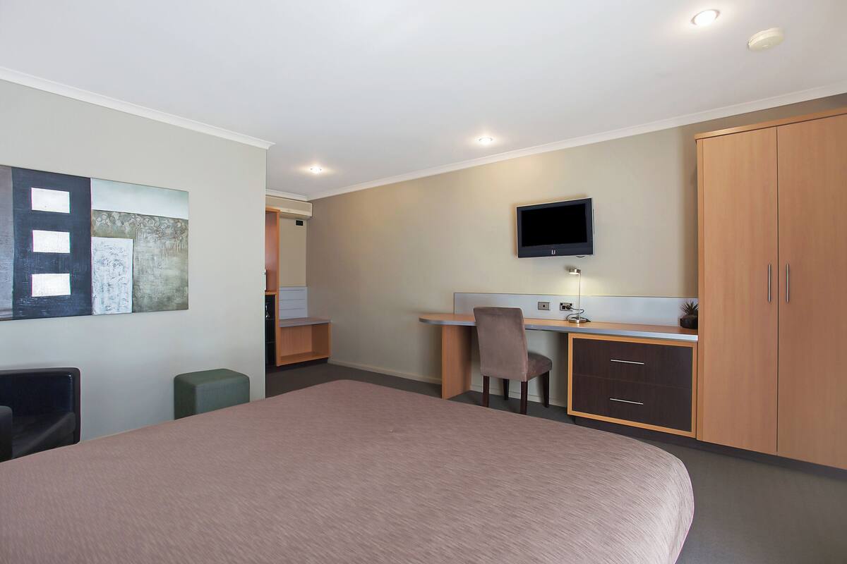 A hotel room with a bed, desk, chair, TV, wardrobe, and artwork on the wall.