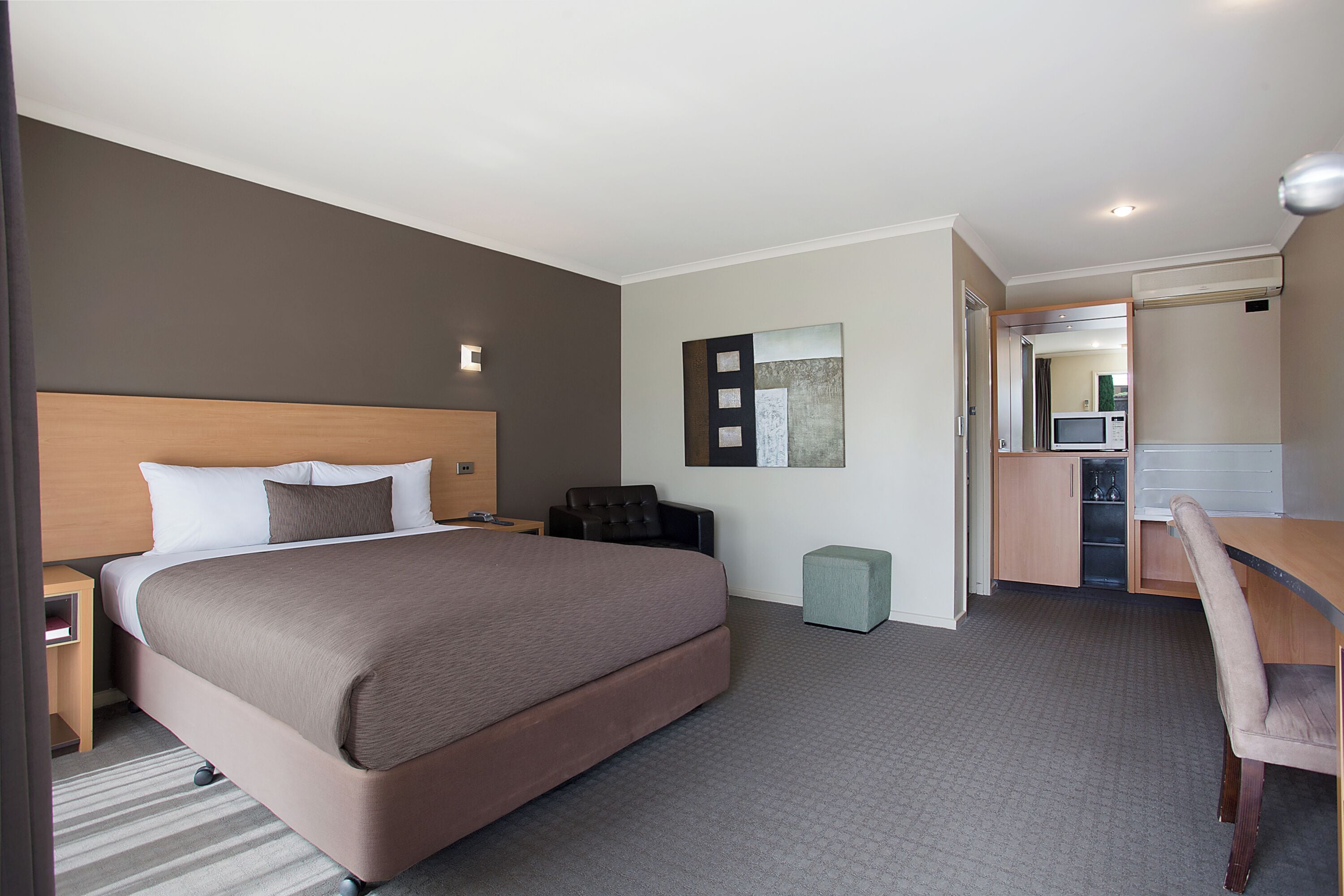 A hotel room with a large bed, a desk, a chair, a small ottoman, a microwave, and a kitchenette.