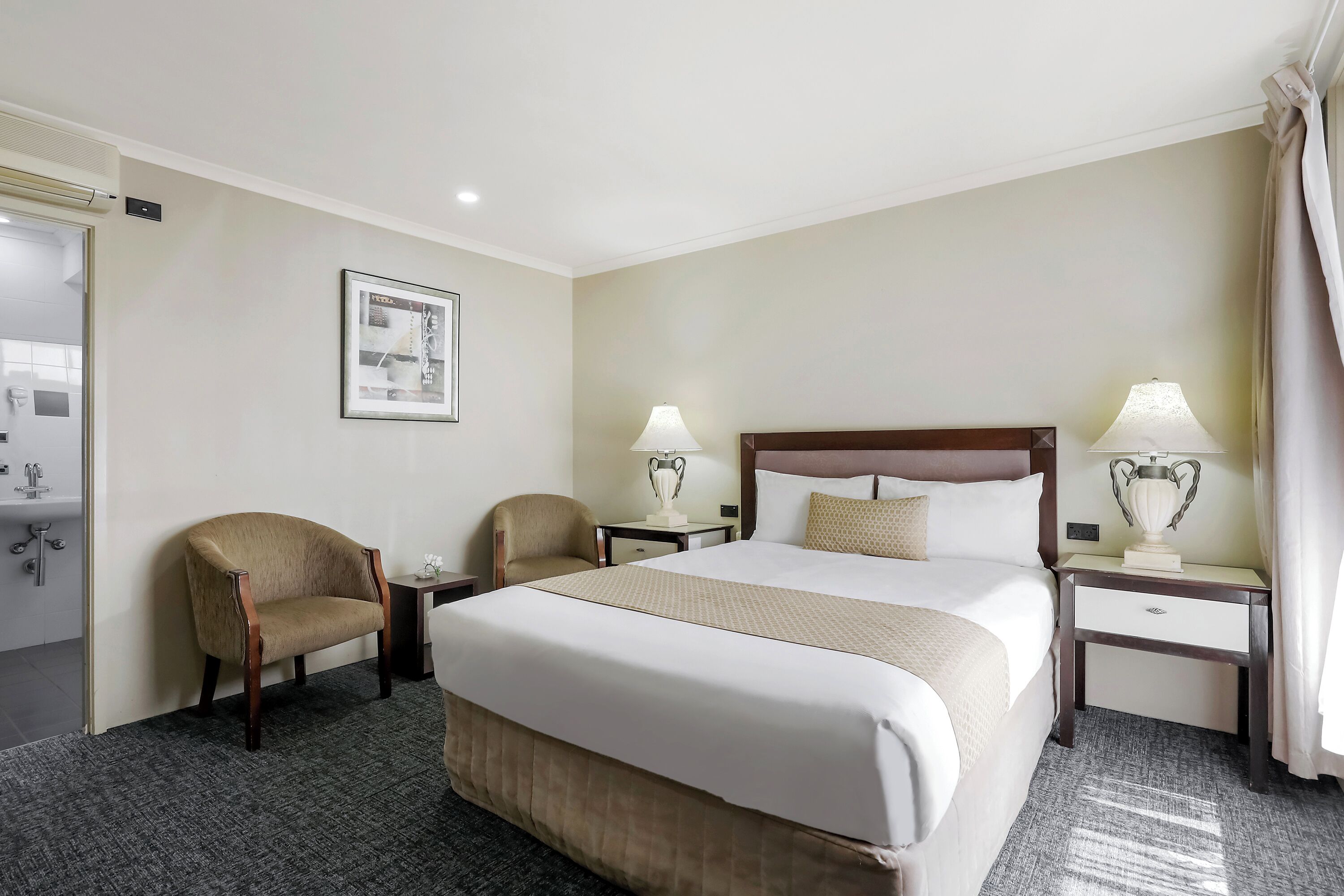 Une chambre d’hôtel avec un grand lit, deux tables de chevet avec des lampes, une chaise, une petite table et un tableau au mur.