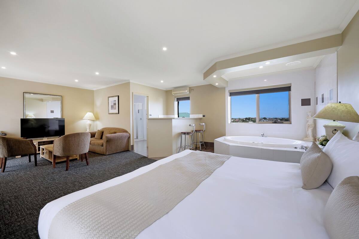 A hotel room with a large bed, a desk with a chair, a sofa, a TV, a bathroom with a bathtub, and a large window with a view.